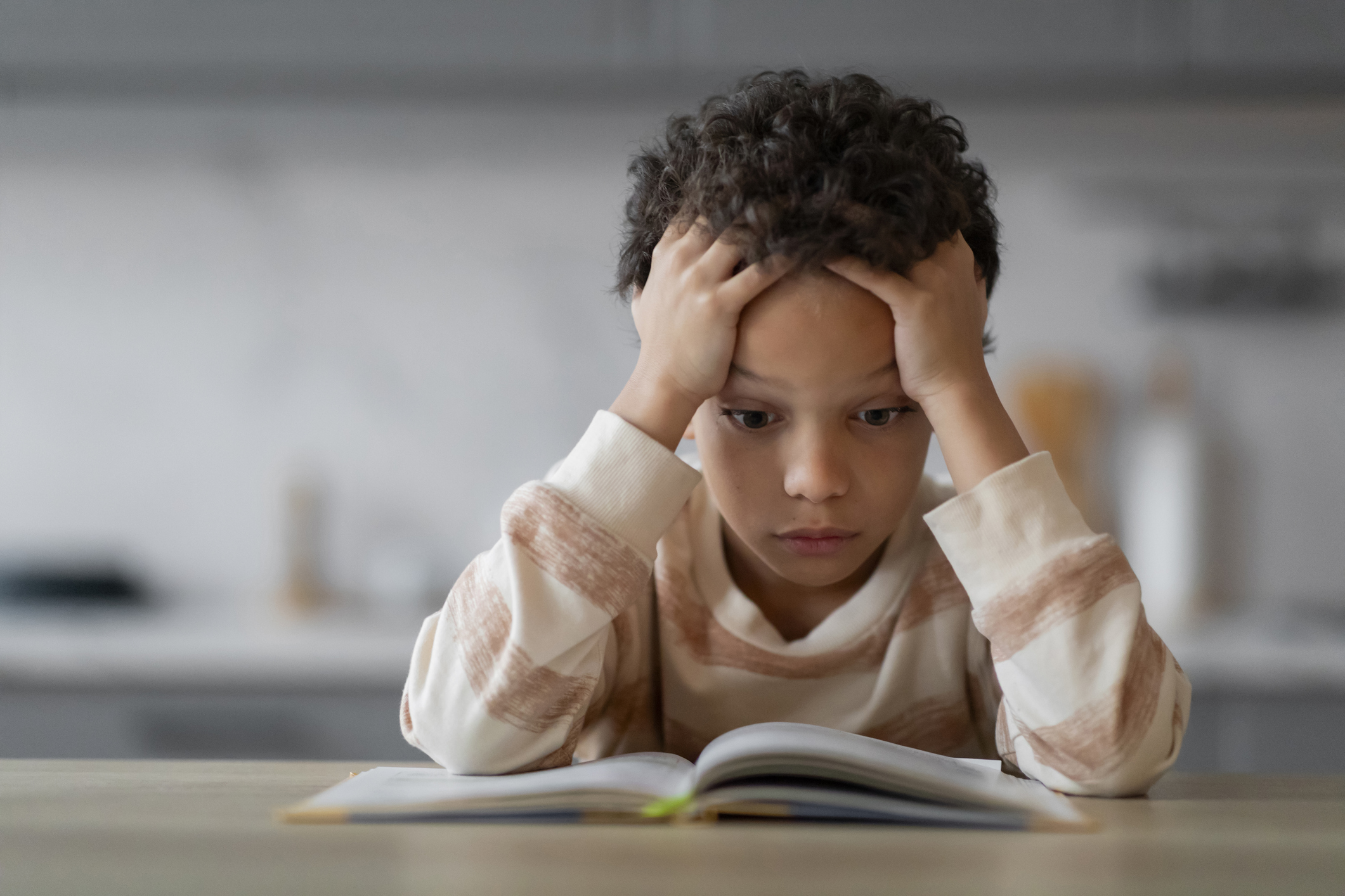 Stressed Little Boy Reading Book At Home And Touching Head